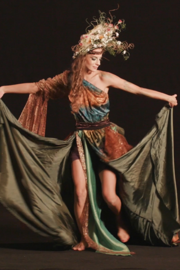 A dancer in ornate costume with a headdress performs, arms extended, against a black background.