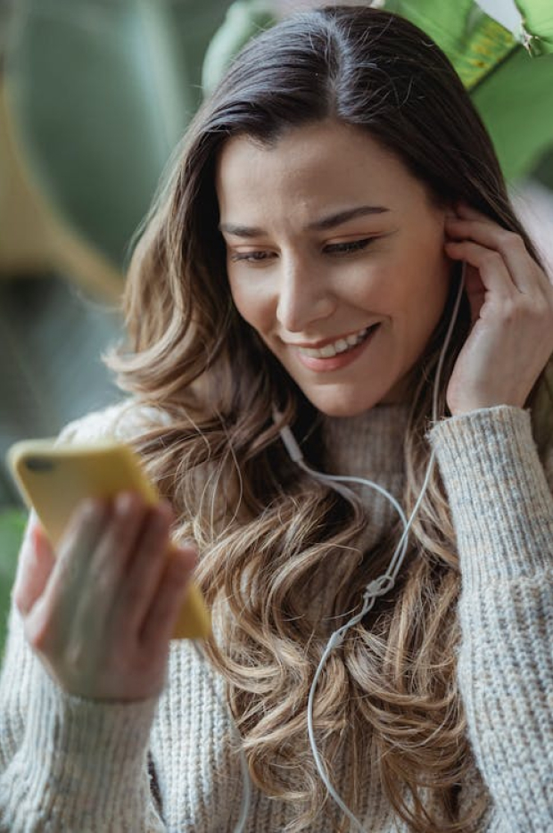A smiling woman with headphones looking at her smartphone, her expression reflecting enjoyment and engagement with the content she is viewing.